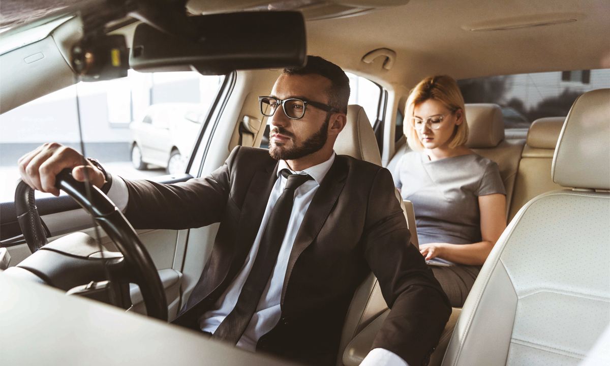 Man in glasses and a suit driving, with a woman in glasses working on a laptop in the back seat