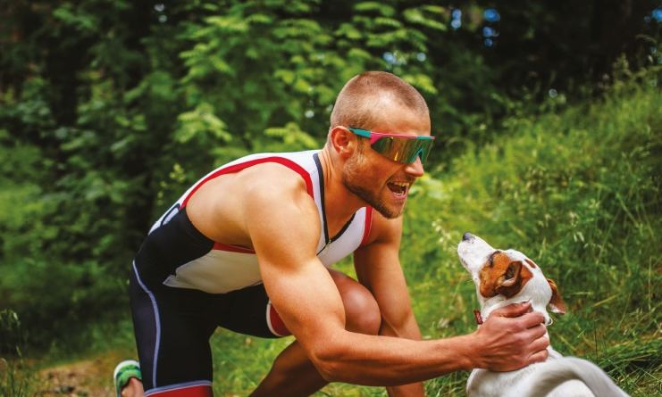 Athlete in sports sunglasses crouching to greet a small dog on a forest trail