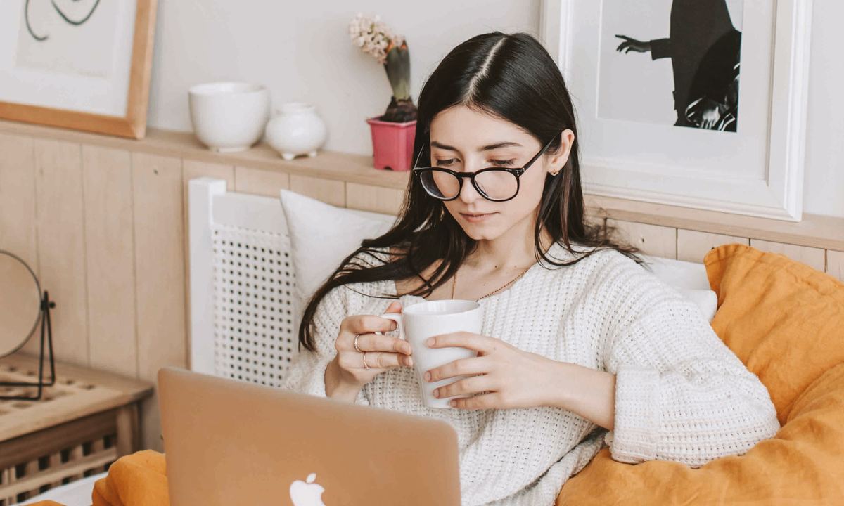 Woman in round glasses holding a mug while working on a laptop in bed